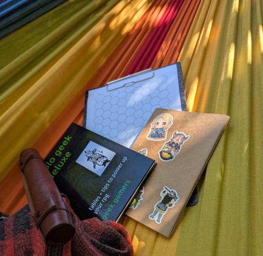 A section of a rainbow hammock, picturing A hexagon sheet on a notepad, a notebook with stickers, a solo gm book and a dice roll