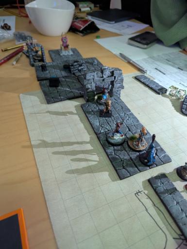 Ein Spieltisch mit Miniatur Figuren und Dungeon tiles, man sieht einen langen Gang der in einen Raum voller Tokens und einer Zauberer Figur mit Hörnern endet. Spielfiguren sind über die Fläche gestellt