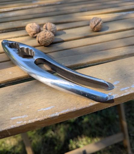 Ein Bildausschnitt auf dem man die Oberfläche eines hölzernen Gartentisches sieht. Darauf ein Edelstahl Nussknacker und vier frische Walnüsse.