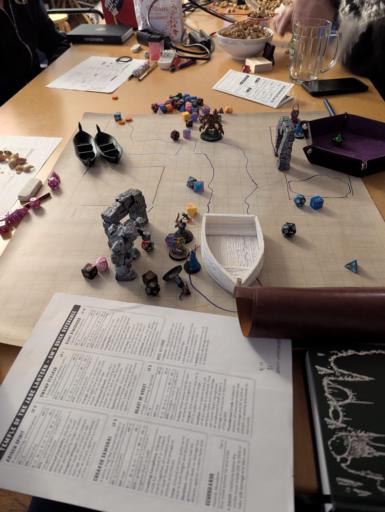 Eine mit Miniaturen und terrain dargestellte Szene auf einer abwaschbaren Spielmatte. Die Gruppe steht an einem Strand vor einem Tor, ein Boot im Rücken. Darum herum liegen Würfel, Regelbücher und Notizen