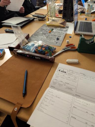 Spieltisch voller Rollenspiel Utensilien. Im Vordergrund ein Cairn Charakter Blatt. Im Hintergrund eine hexmap von hot spring Island