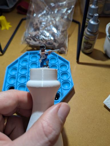 Picture of a half painted resin miniature on a 3d printed painting handle. Painting tools and a make shift palette in the background