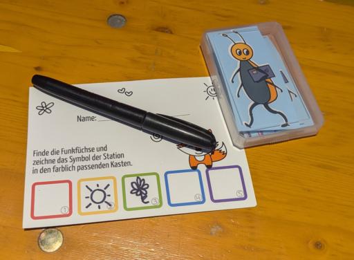 Eine Postkarte mit dem Aufdruck: finde die Funkfüchse und zeichne das Symbol der Station in den passenden Kasten. Zwei von fünf Kästen sind schon ausgefüllt. Daneben liegt eine Spielkarten box mit einem Sticker der einen comic haften Käfer zeigt